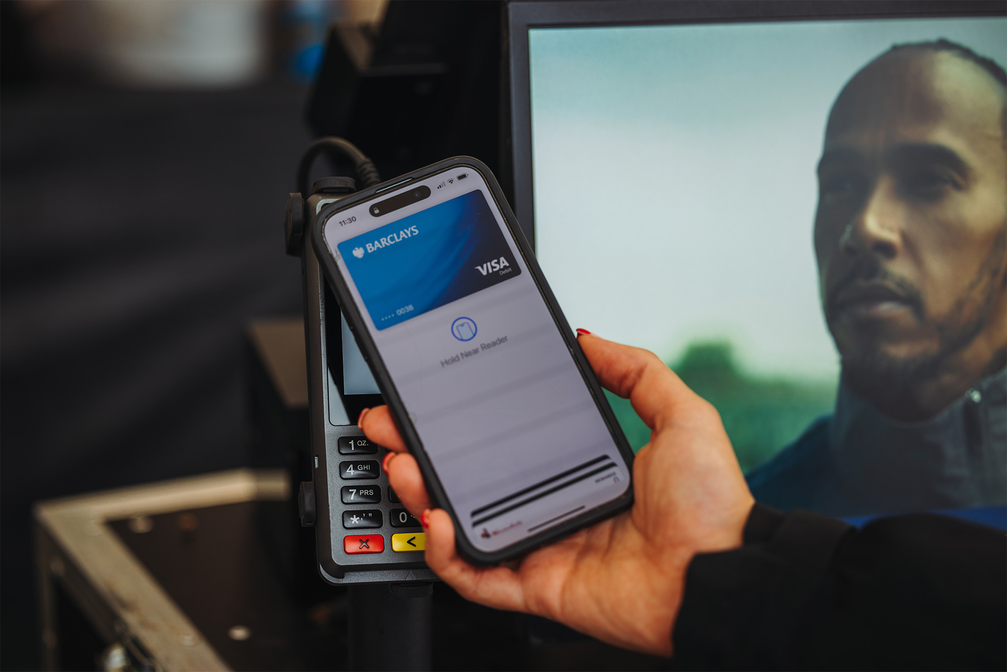 A person holds a smartphone displaying a digital wallet with a Visa card from Barclays, ready to make a contactless payment on a card reader. A screen in the background shows a mans face partially visible.
