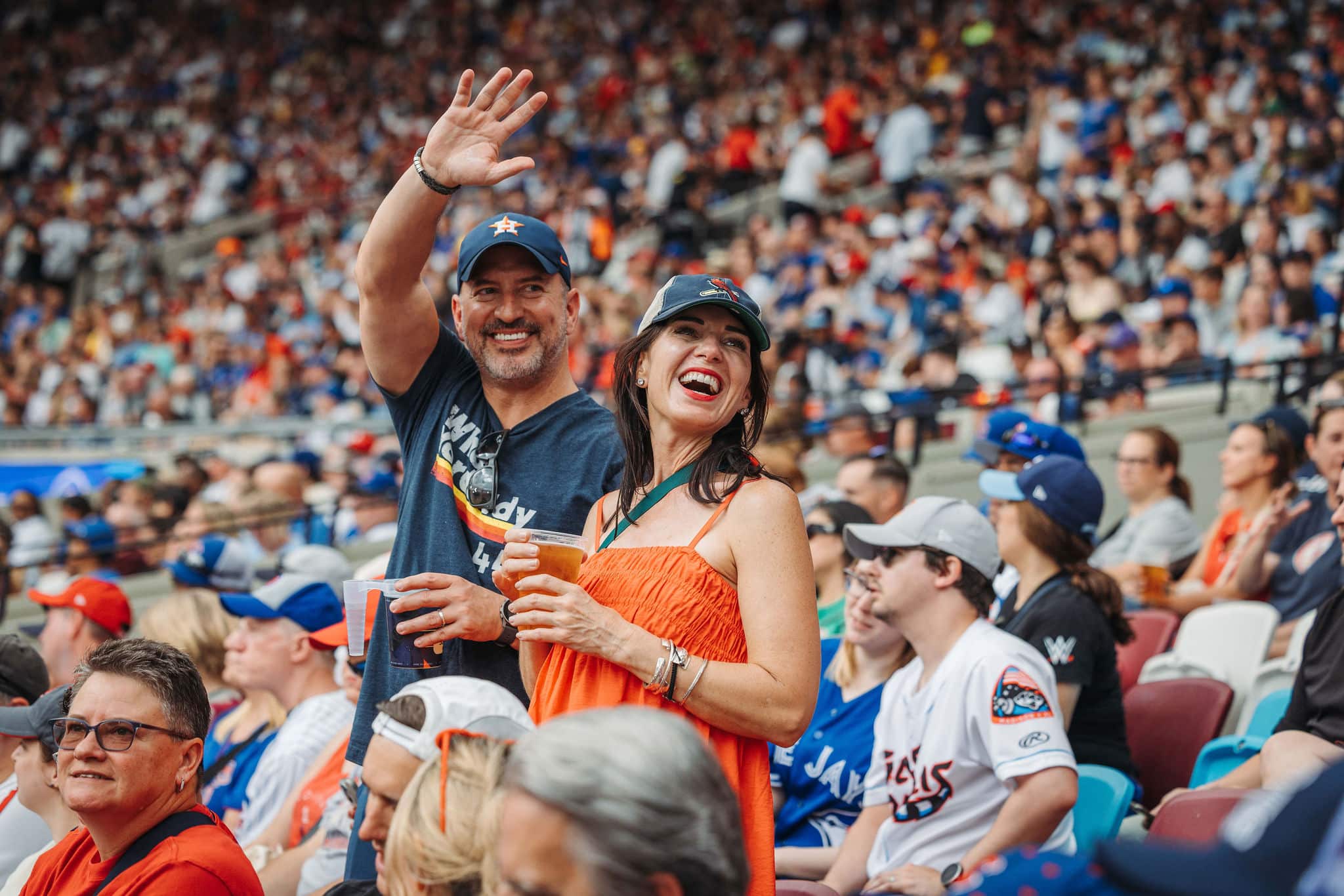 A man and woman stand in a crowded stadium, cheering and waving. The man wears a baseball cap and holds a drink, while the woman in an orange dress smiles broadly. Spectators around them enjoy the game.