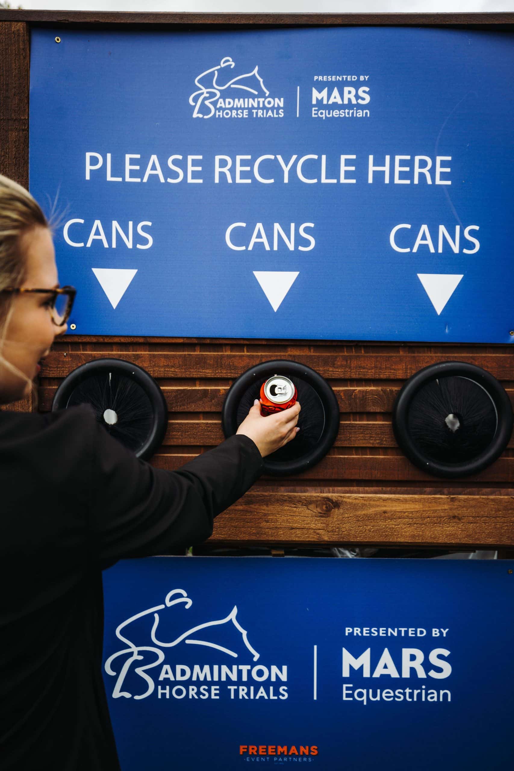 A person recycles a can by placing it into a designated recycling bin at an event, with a blue sign above reading PLEASE RECYCLE HERE and CANS at the Badminton Horse Trials.