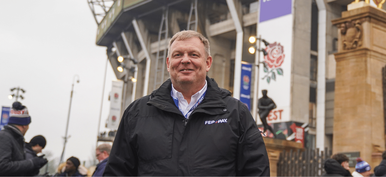 A man in a black FEP-PAY jacket stands smiling outside a stadium with banners, statues, and people in the background on a cloudy day.