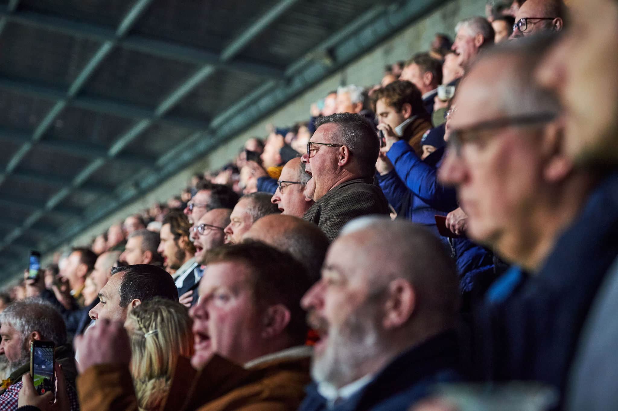 A crowd of spectators, mostly men, watch intently from stadium seats, with some cheering and others focused on the action. The background shows the stadium roof structure above them.