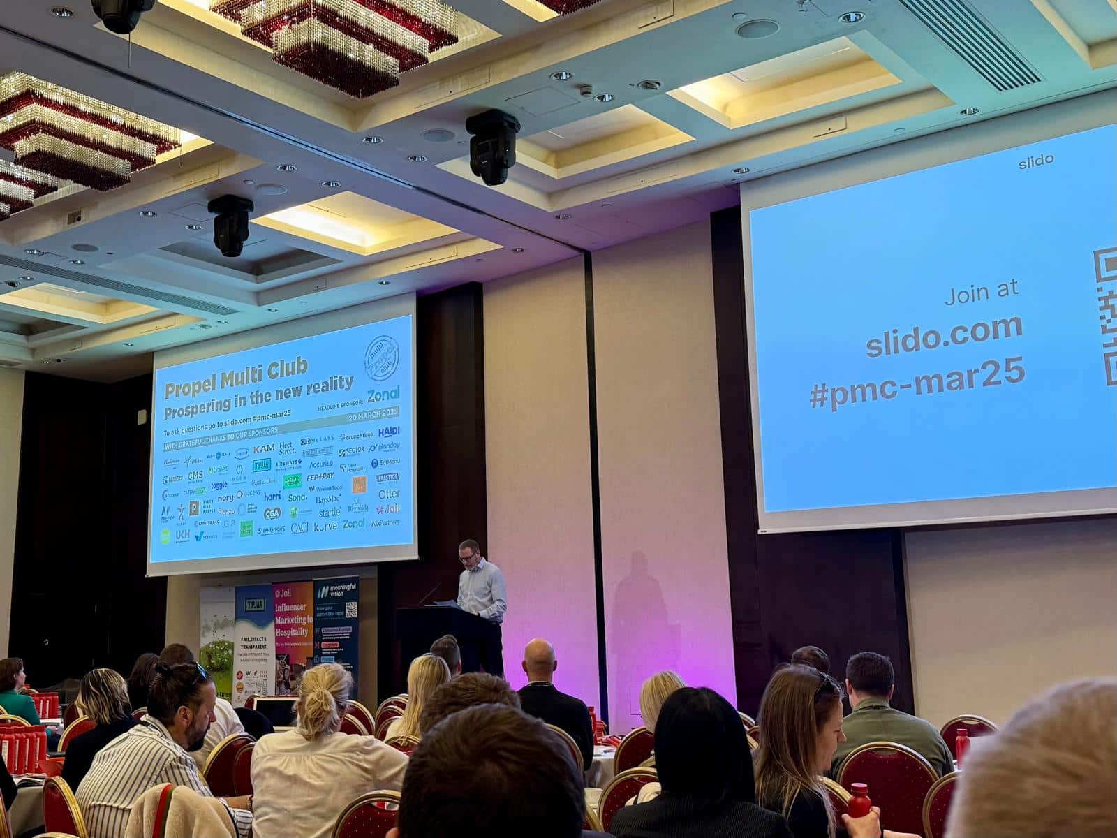A speaker presents at a conference in a room with rows of seated attendees. A large screen displays text including Propel Multi Club and hashtags. Red cushioned chairs arranged in rows are visible.