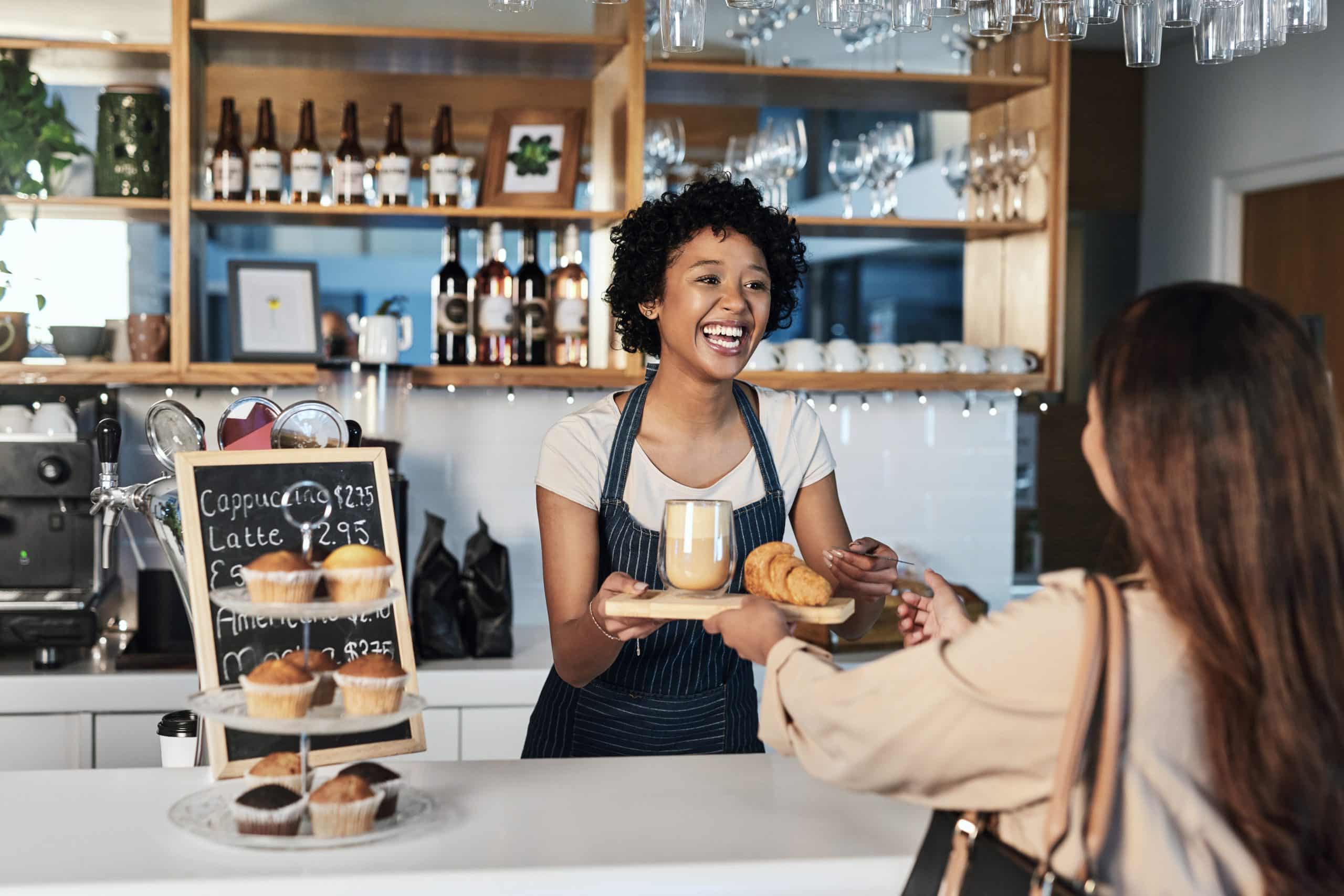 A friendly barista smiles while handing a tray with a coffee and croissant to a customer at a modern café counter. Muffins are displayed beside a chalkboard menu, and wine glasses hang above the counter.